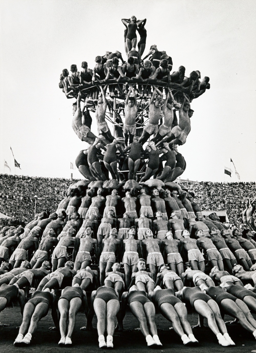 Black and white photograph of athletic men and women holding positions to create a human pyramid several stories high in the center of a packed amphitheater full of people.