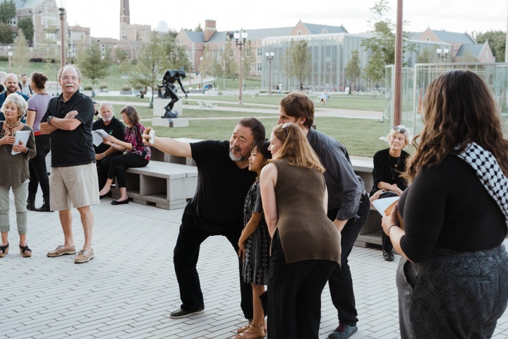 Artist Ai Weiwei takes a selfie with a few attendees at his exhibition opening. Everyone is gathered outside, in front of the green park area outside the Kemper Art Museum; other onlookers and patrons mingle.