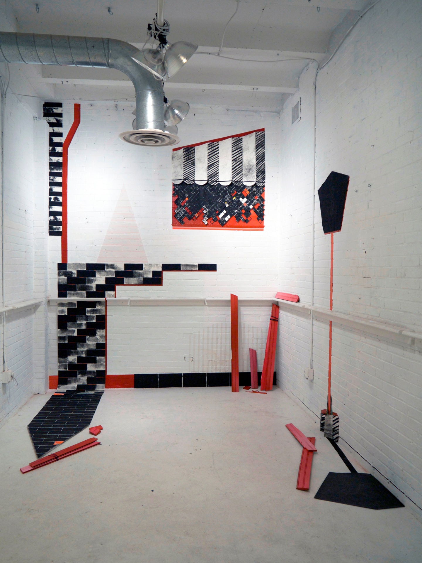 Against white brick walls and a white concrete floor, an installation: a black and white pattern tiled across some of the bricks in the wall lead to the upper edge of the wall; red tiles/material outlines some of the patterns, a few pieces of the red material also stacked together in the foreground.