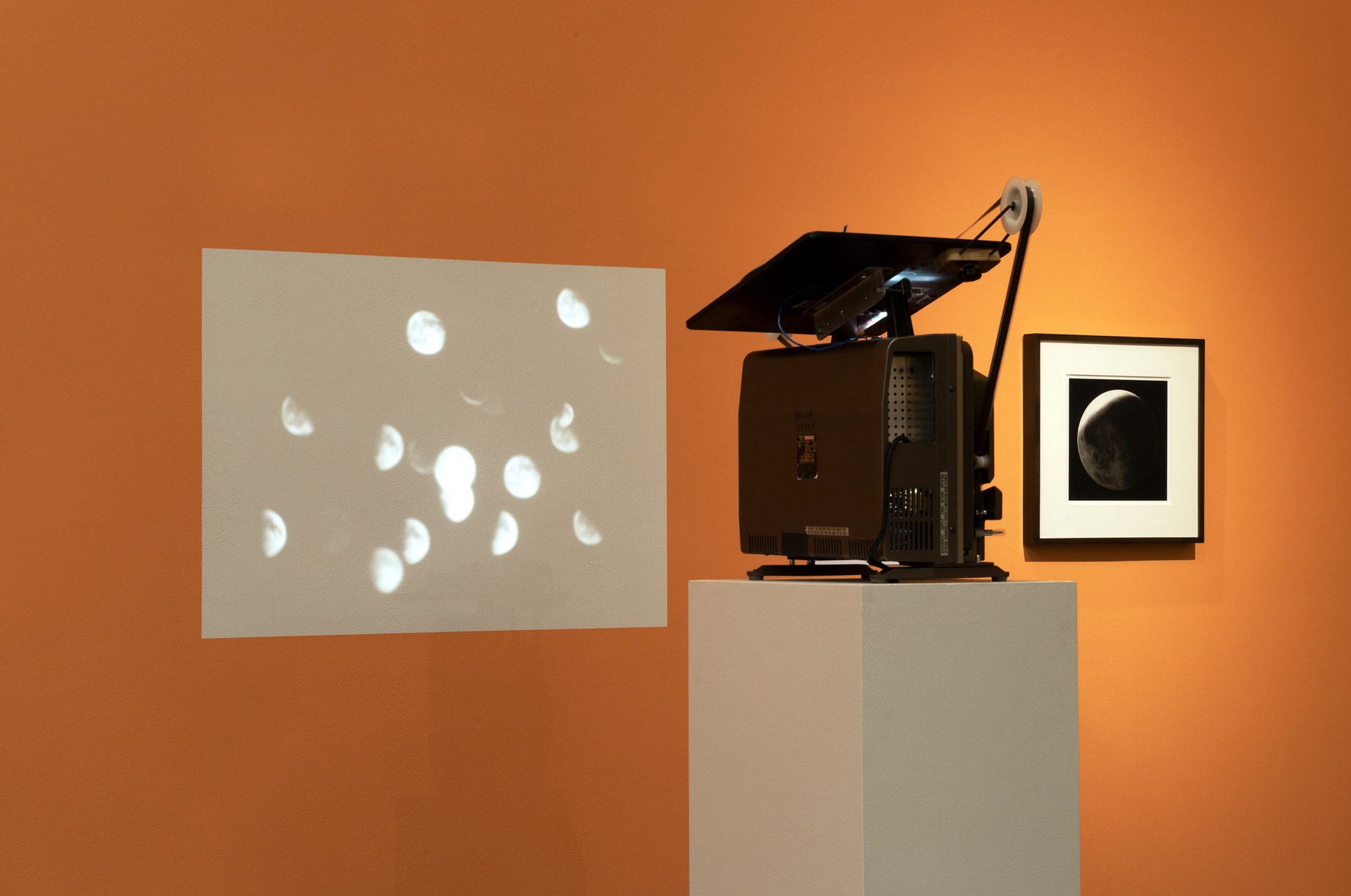 A projector sits on a gray pedestal and projects an image of a cluster of full, half, and crescent moons onto an orange wall, next to a framed photograph.