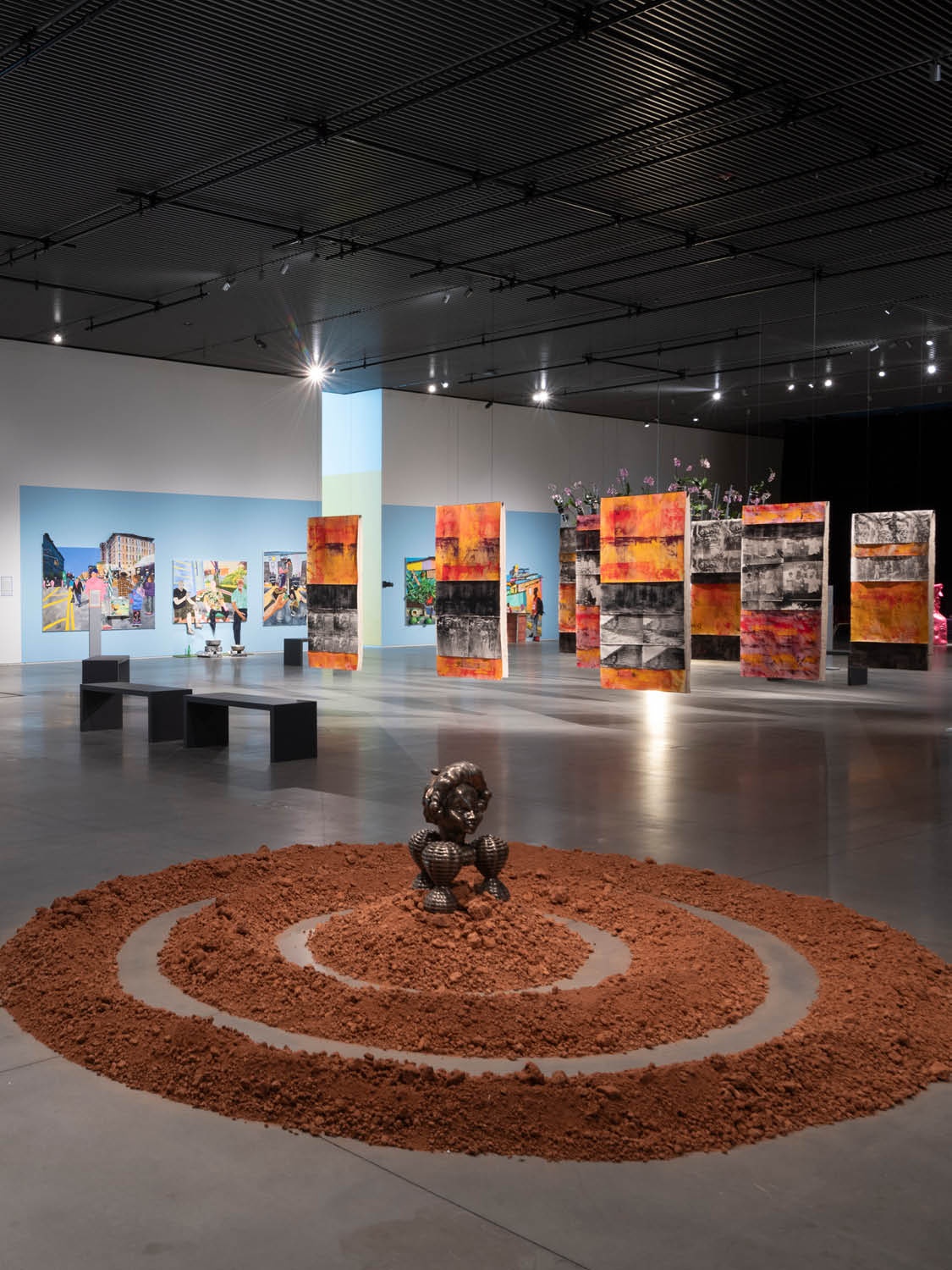 A view of an exhibition in a large art gallery without any walls. In the foreground is an artwork on the floor that consists of three concentric rings of red dirt. At the center sits a bronze sculpture inspired by the Benin Bronzes, historical artworks from Benin, present-day Nigeria. In the background are other paintings and installations. In the background are various paintings hanging from the ceiling and walls.