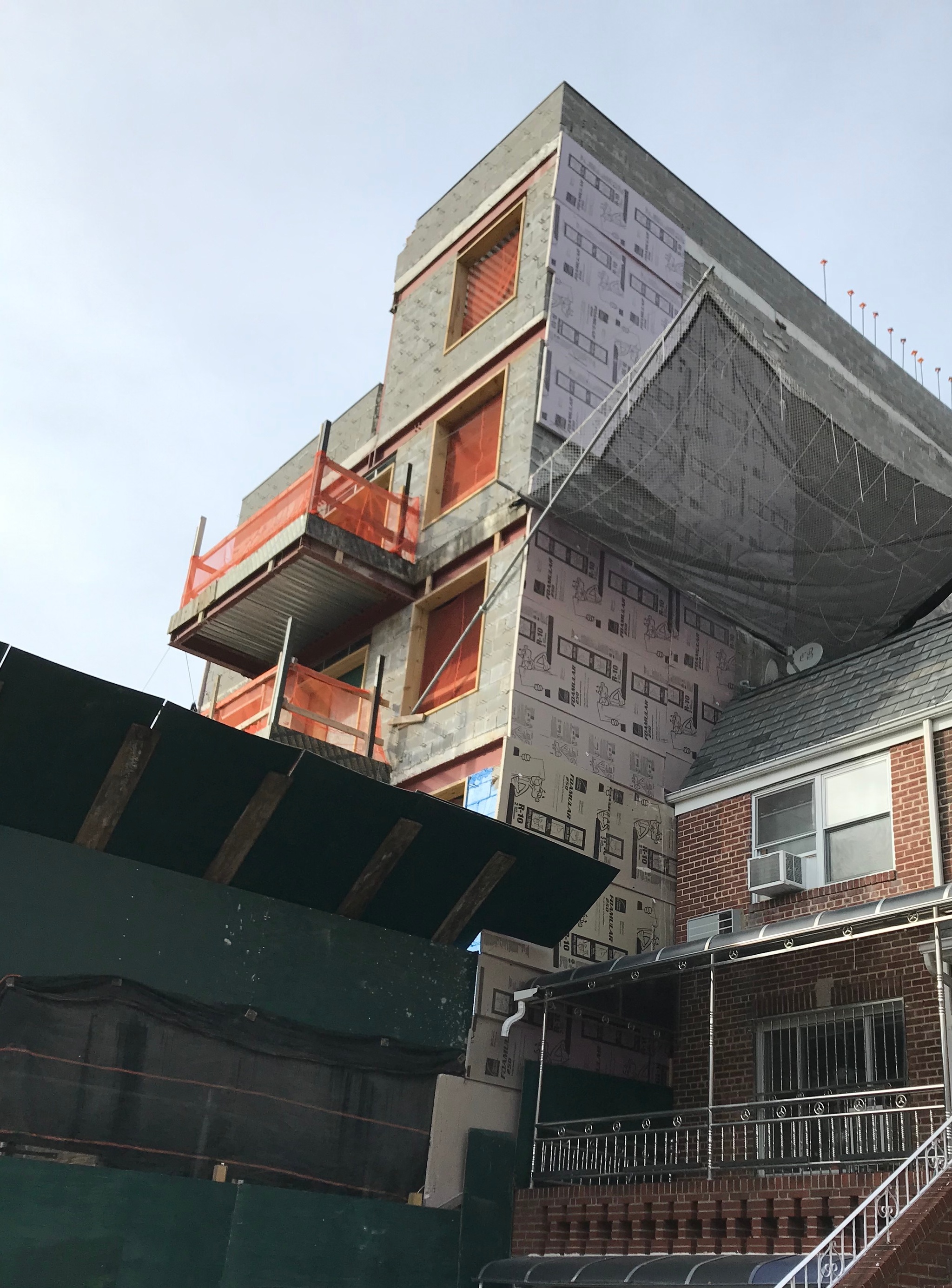 A five-story apartment building under construction. It has unfinished concrete walls and orange plastic netting encloses the spaces where windows and balconies will be. To its right, a smaller two-story brick house sits beside it. The house has steel railings along its front porch. 