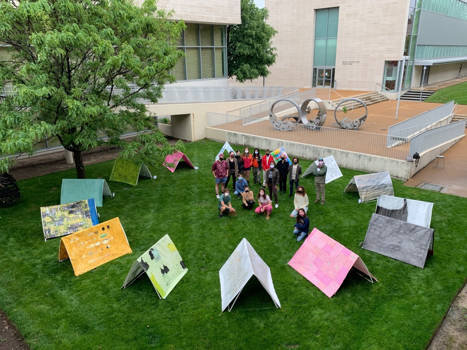 People stand next to different colored and patterned triangular tents. The tents and people are arranged in a ring on a grass field, surrounded by plazas and buildings. The people are wearing masks. 