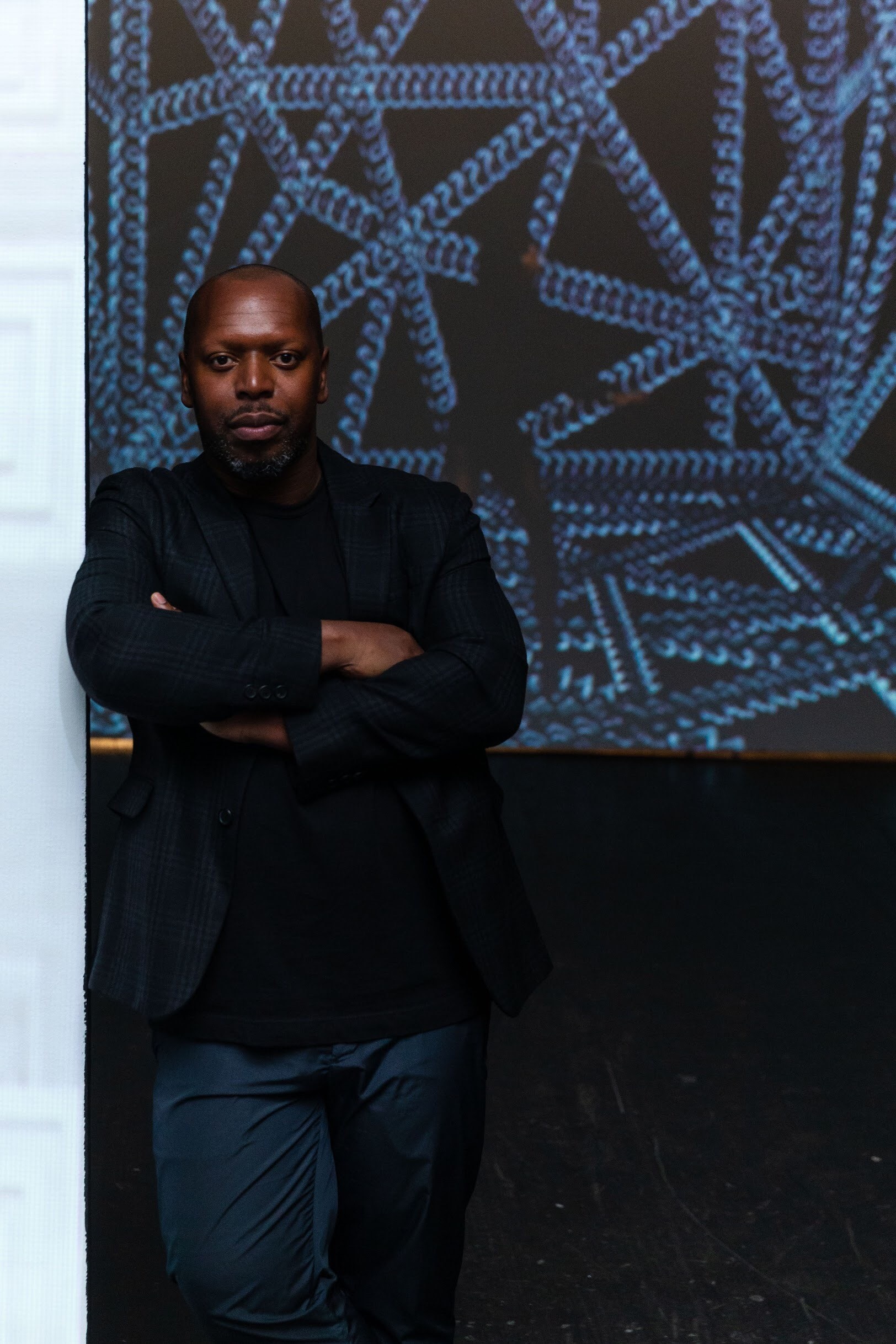 An portrait of the artist Rashaad Newsome standing in front of a digital projection with his arms crossed across his chest