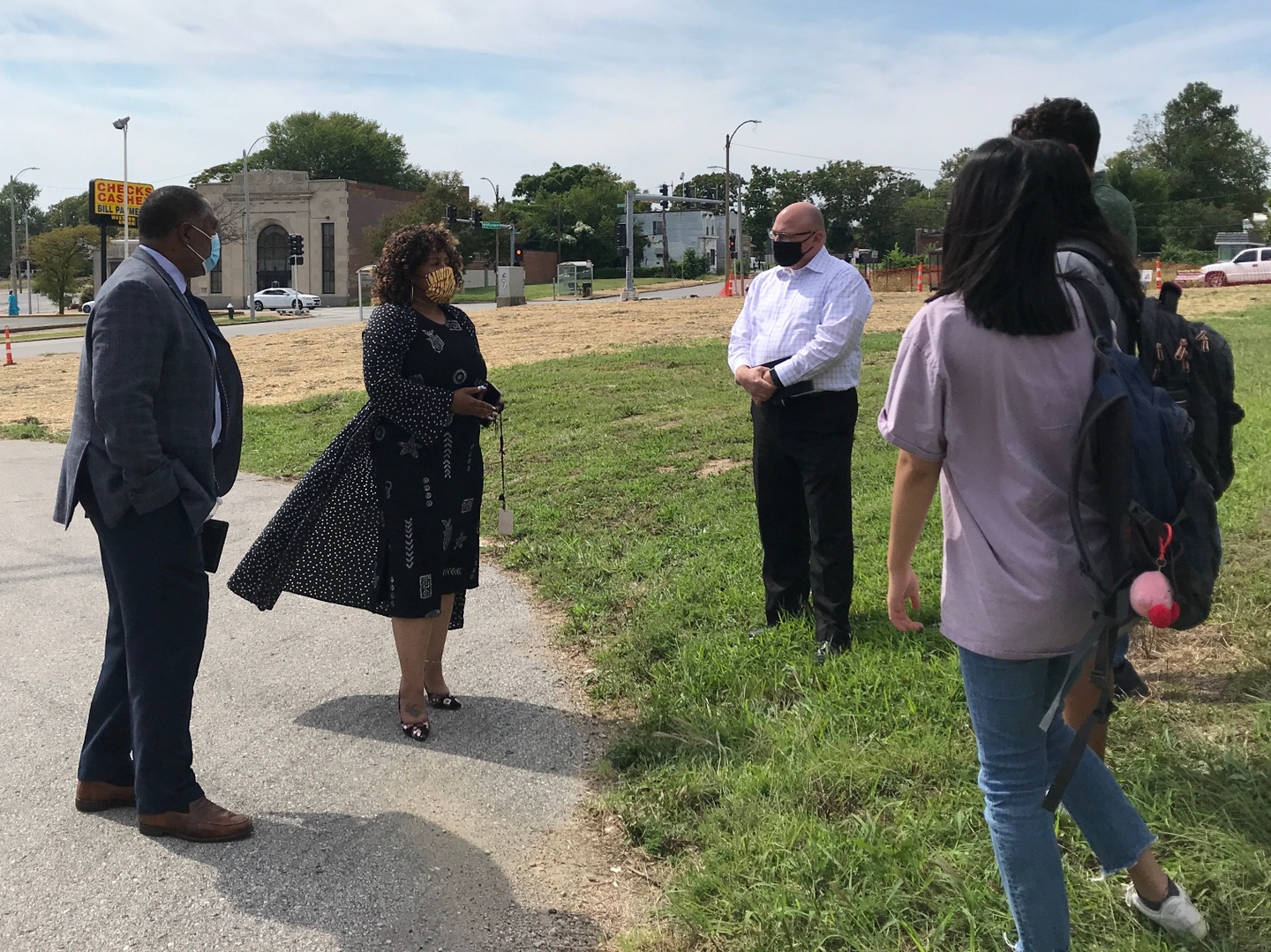 A group of people stand in a grass field and on a sidewalk. Behind them is a street. 