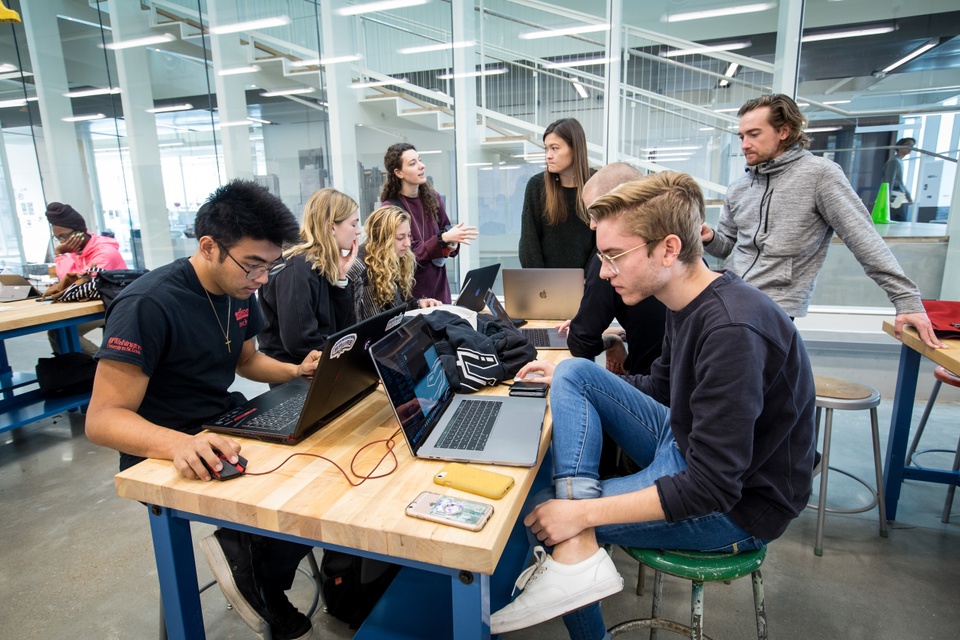 Group of students work on laptops at a table in the Caleres Fabrication Lab while instructors advise.