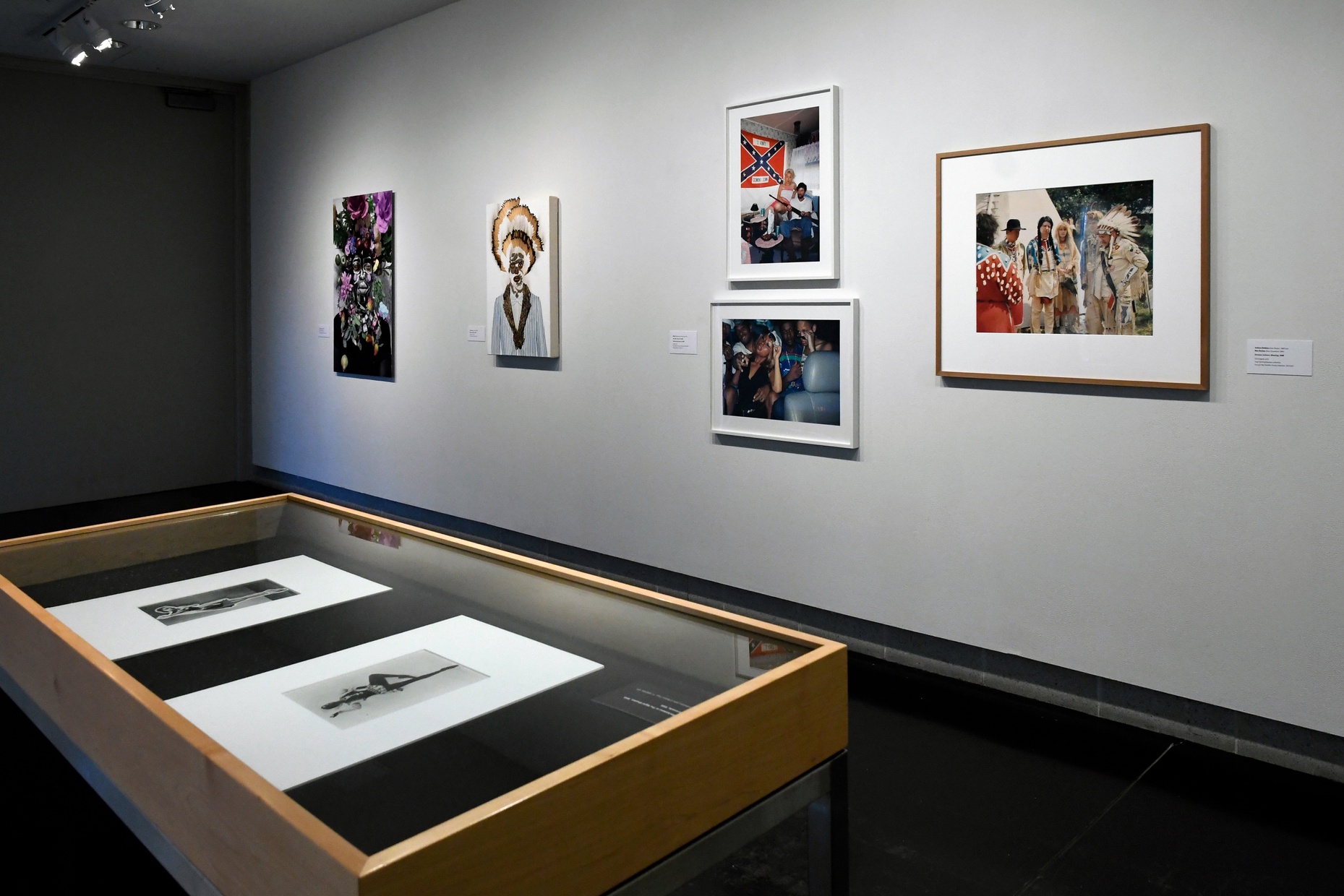 Five photographs of people hang on a gray wall with a case holding two black and white photographs visible in the foreground.