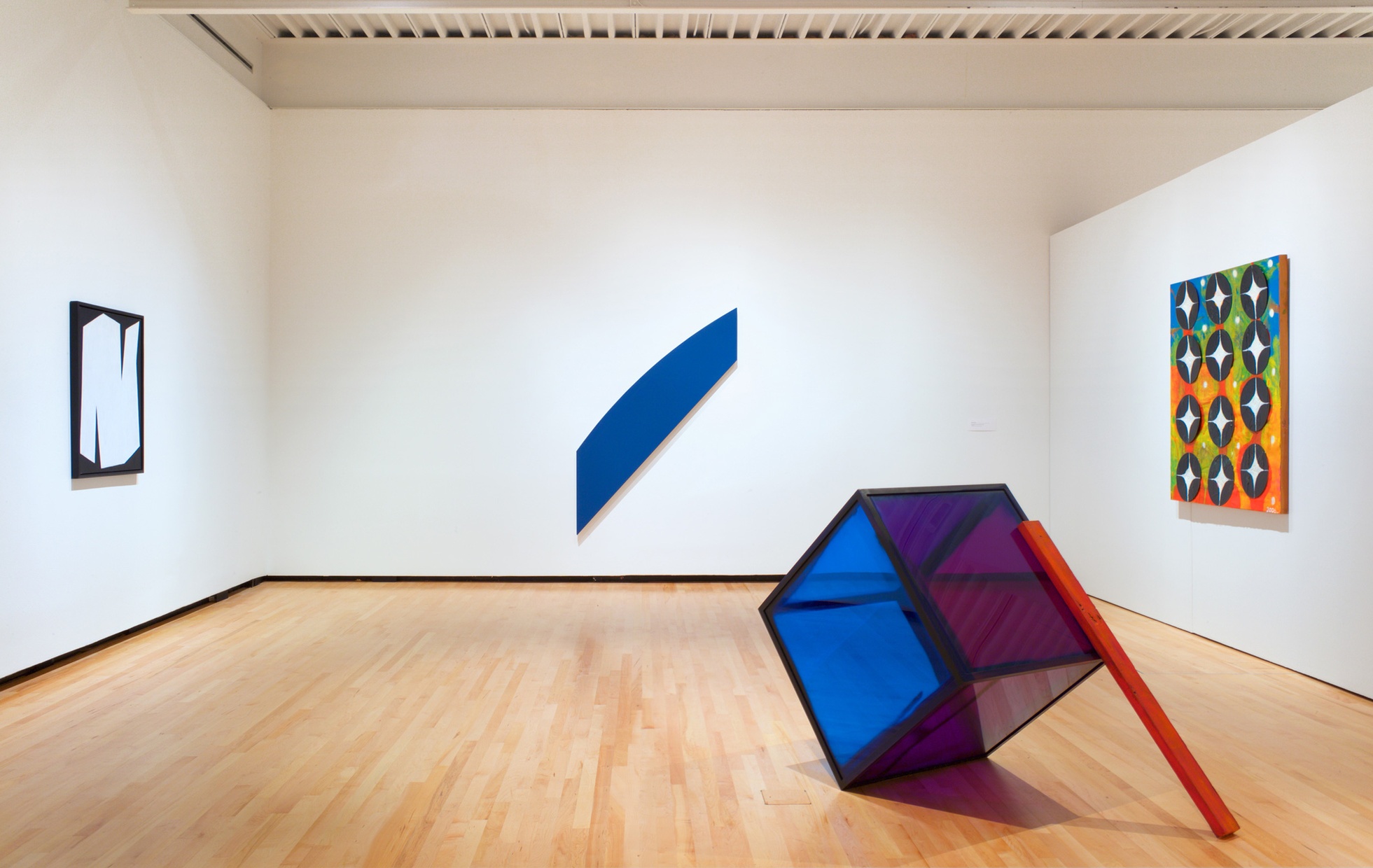 A white-walled room with an abstract painting on each wall and in the center is a purple and blue cube tilted at an angle with a red stick leaning against it. 