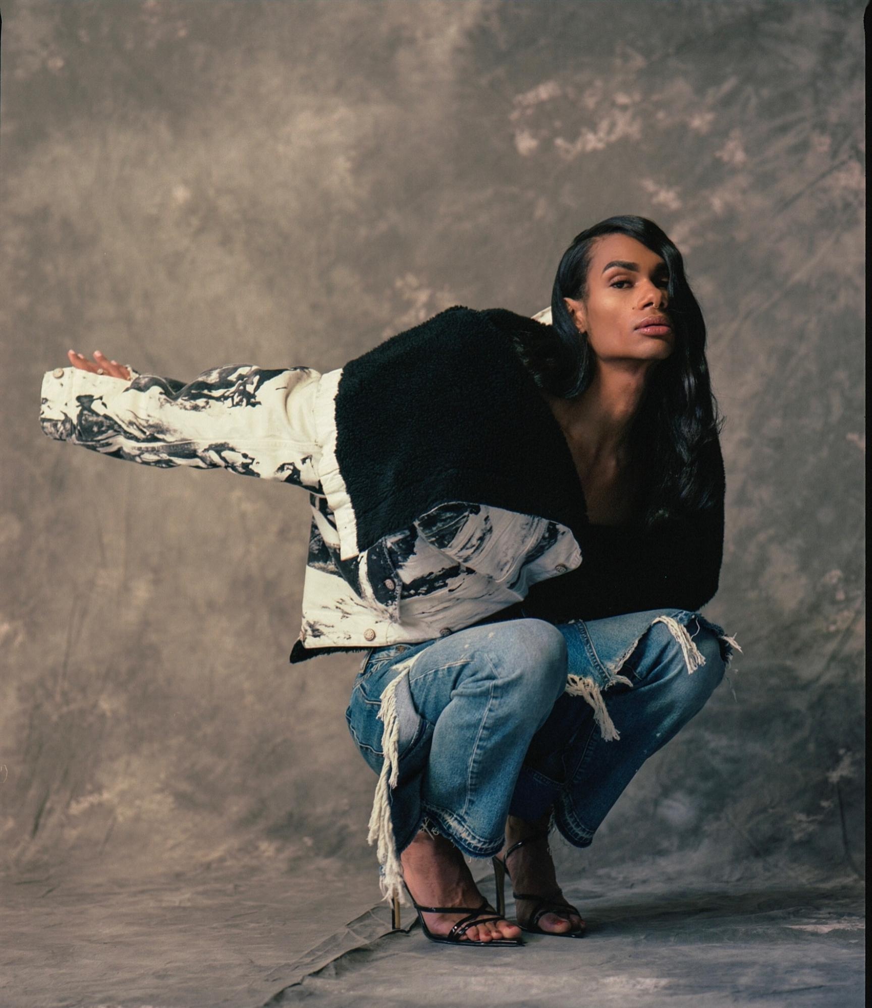 A portrait of artist z tye crouching in heels and a pair of jeans against a studio backdrop with arms outstretched behind her. Her black, straightened, wavy hair falls over one eye.