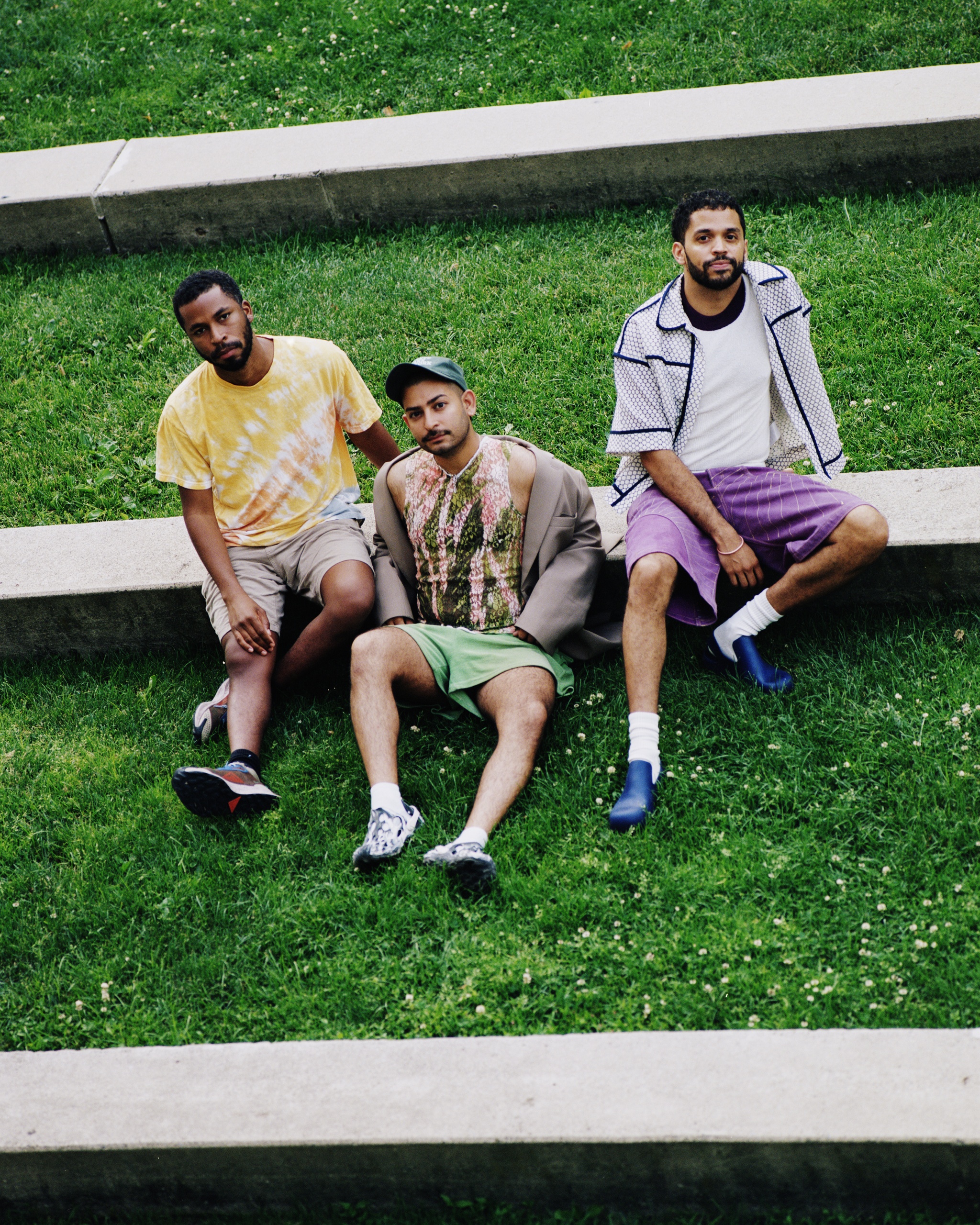 Three members of the art collective Papi Juice, all people of color, lounge on a grassy amphitheatre-like structure. They wear casual summery clothes: tshirts, shorts, baseball caps. 