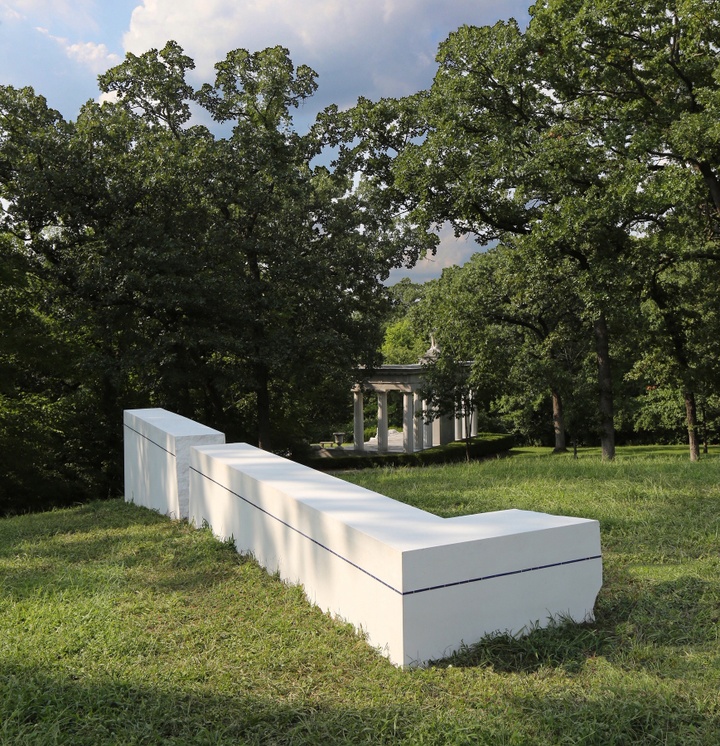 An 'L' shaped white concrete sculpture with a dark blue line on its sides on a grassy plain