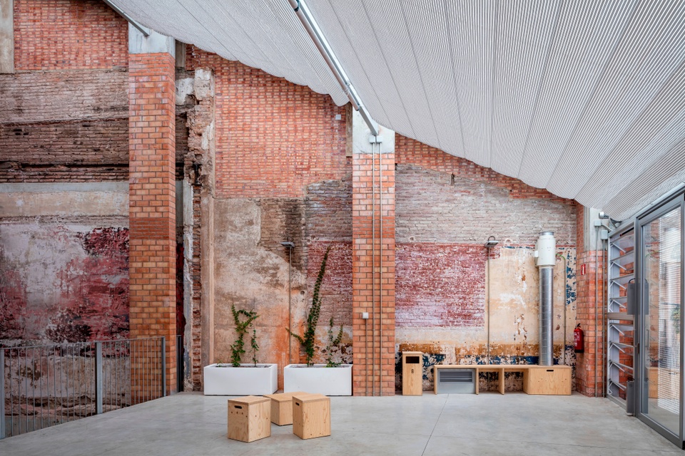 Interior building view, showing a slatned, metallic ceiling plus walls revealing different types and colors of distressed brick. On the concrete floor are several small wooden pedestals and a bench on the far wall, plus two white planters with plants in them.