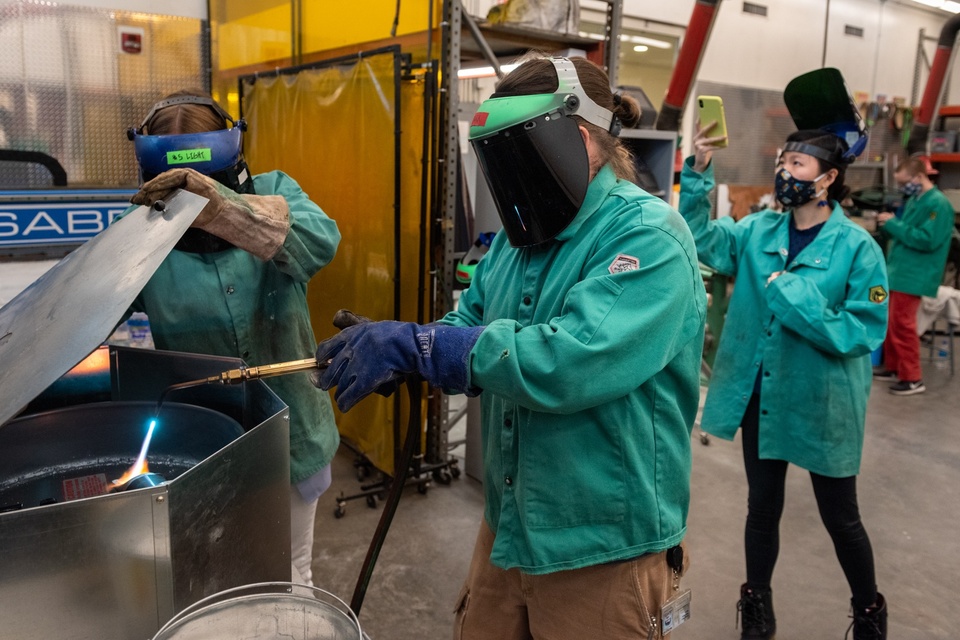 Two people in PPE melt bronze with a handheld torch in a crucible.
