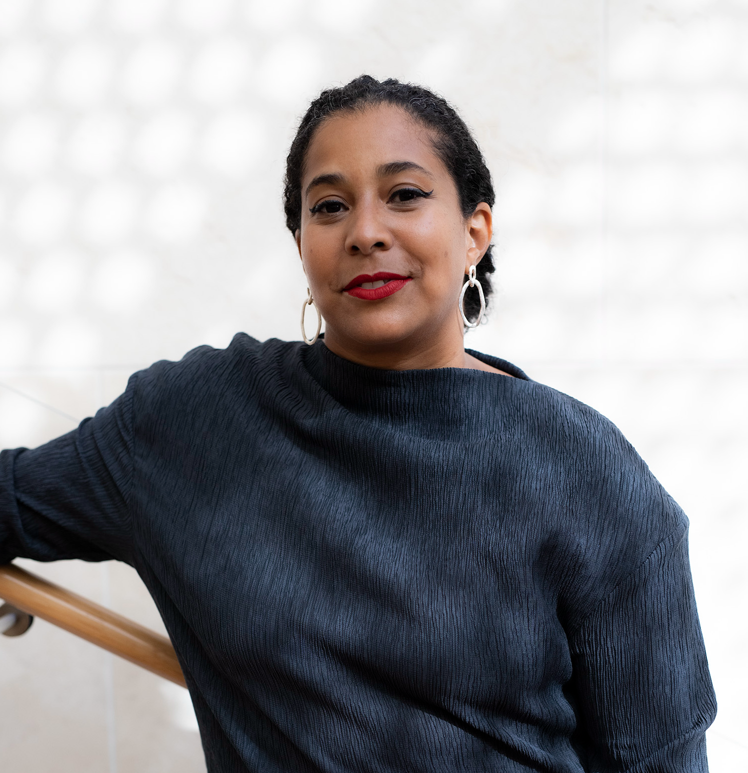 A portrait of Vivian Crockett who leans against a stairwell banister against a white wall. She wears a navy blue sweater, red lipstick, and has dark hair pulled back behind her head. Photo by Ciara Elle Bryant