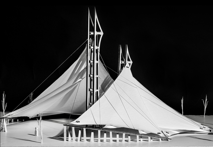 Black-and-white photo of an architectural model, featuring two triangular, sail-like coverings hung from vertical structures, each with two points.