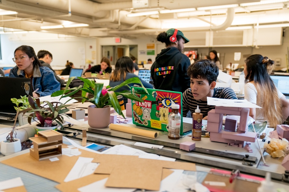 Large group of students work on models and laptops surrounded by model-making supplies in an open format work area.