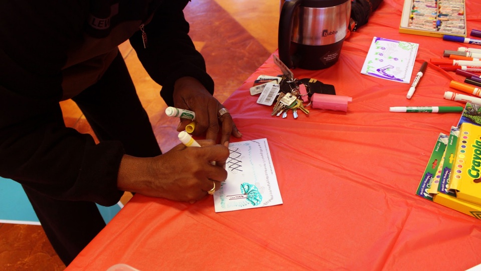 A person holding a marker writes on a postcard on a table with other markers and postcard supplies.