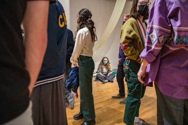 A crowd parts to reveal a person sitting on the floor next to a sculptural installation, listening to headphones.