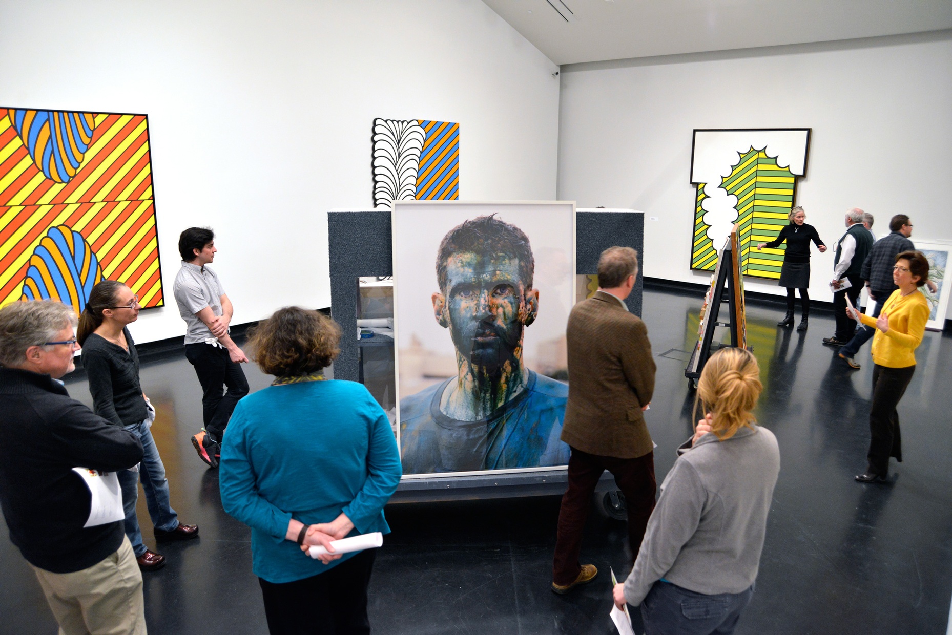 A group of people scattered throughout a gallery space with large, colorful, abstract prints on the walls and a large photograph of a young, white man with blue paint on his face sitting in the center of the gallery.