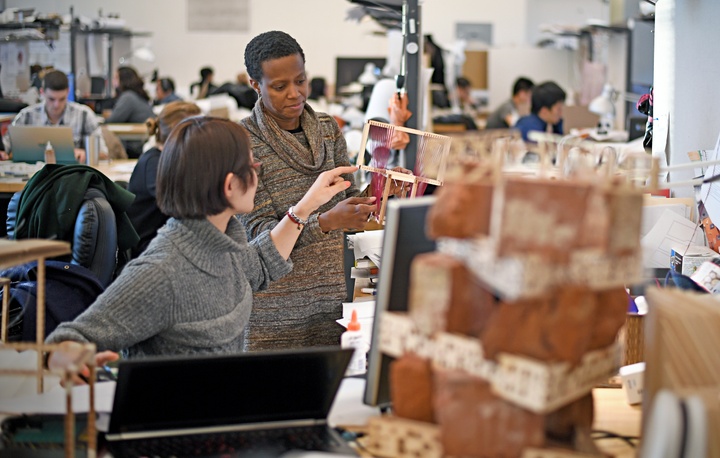 Deep studio space with dozens of students and semi-private desks piled high with models.