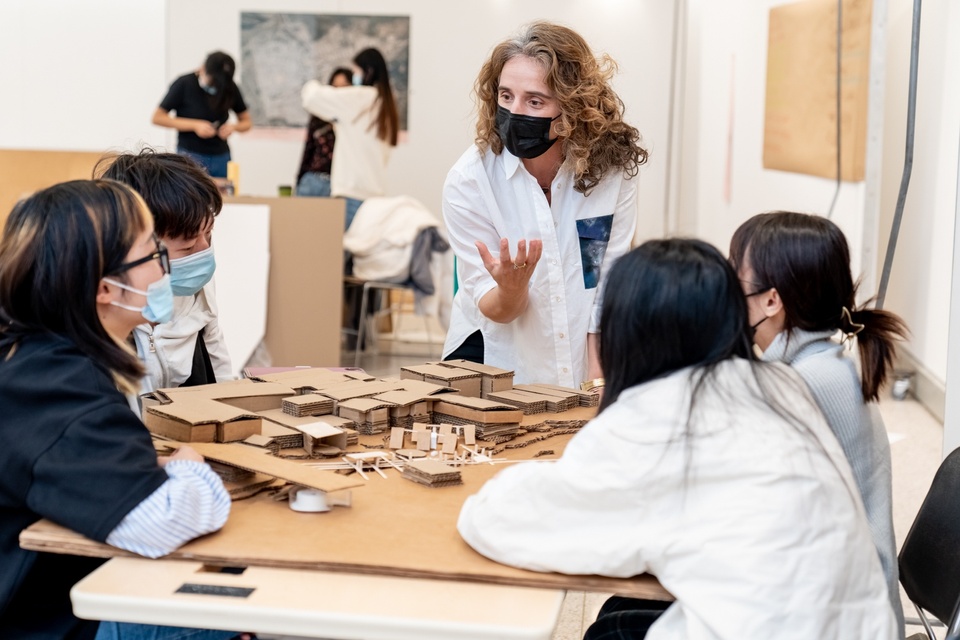 Instructor speaks enthusiastically to students seated around a table holding a cardboard city plan model under construction.