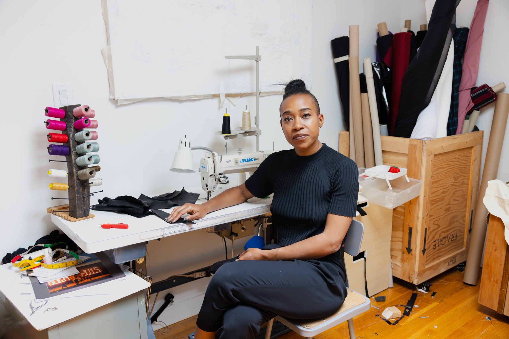 Artist Calli Roche, a Black person with hair pulled back in a tight bun, sits at a sewing table in their art studio. They are turned out to look directly at us and appear relaxed with one arm on the table. Around them is a sewing machine, textiles on tubes leaning in a corner, and skeins of pink thread on a carousel.