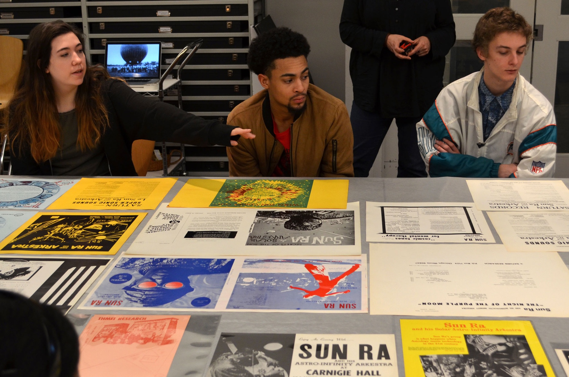 Three students sit behind a table covered in colorful prints and album covers.