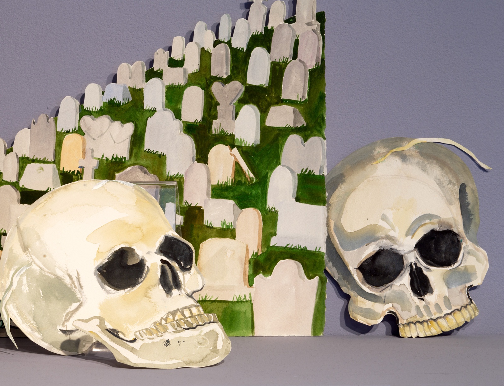 Painted paper cutouts of two skulls lean against a painted cemetery scene, all against a light purple wall.