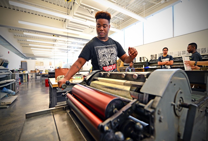 A person stands behind a letterpress machine, turning the crank.