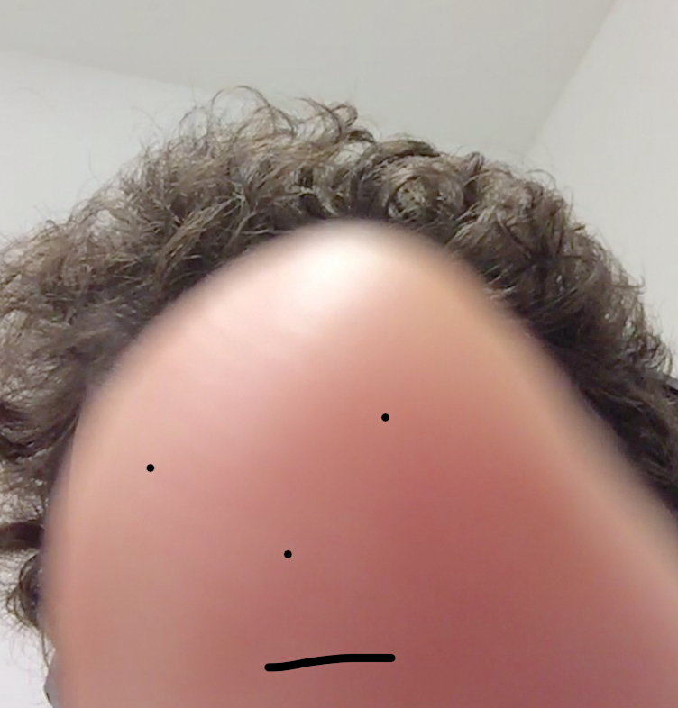 A photo of a white person's fingertip with a face drawn on it and photographed in front of person's head so it looks like the finger has short curly, brown hair