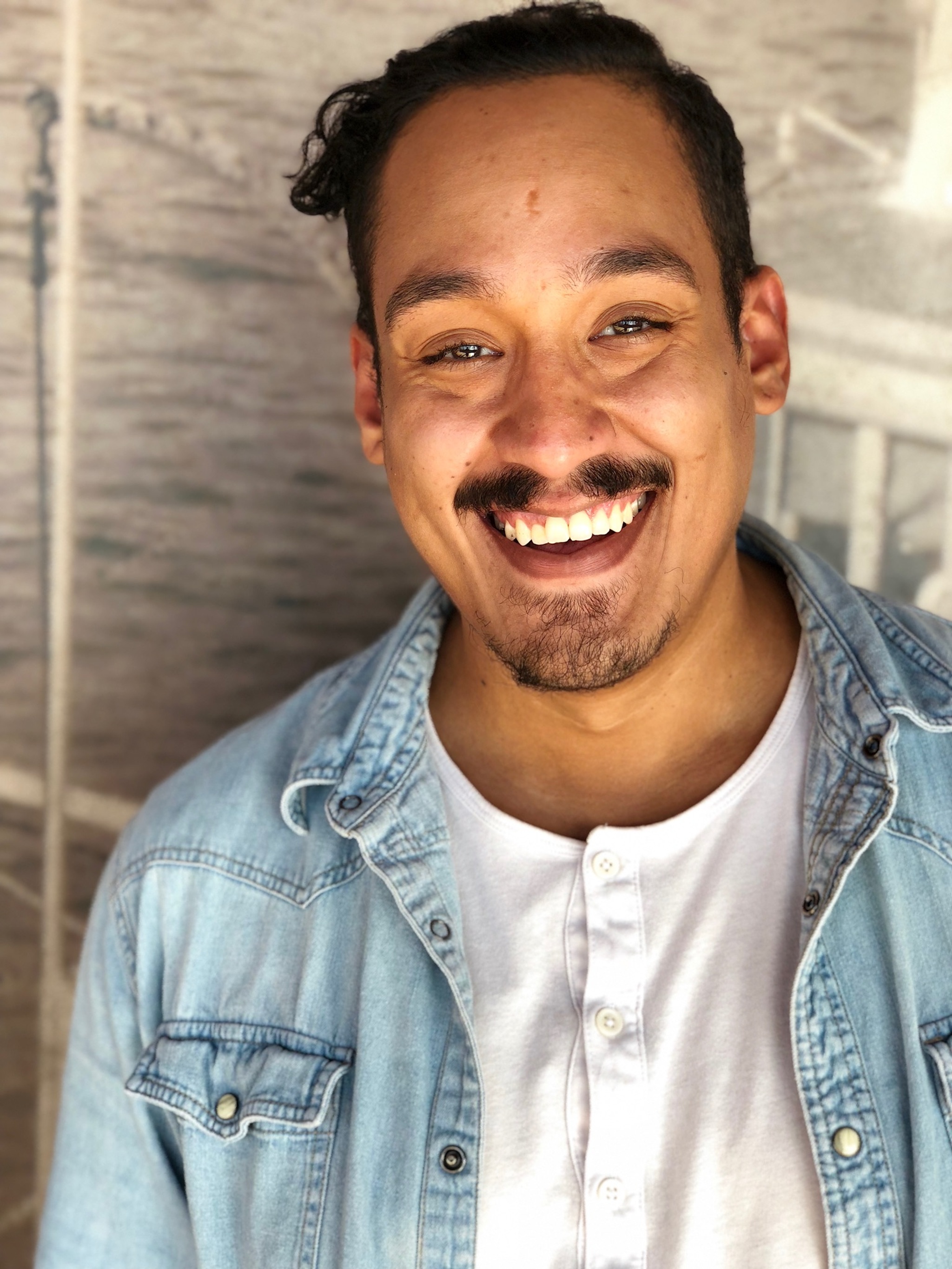 A man smile broadly in a space lit so that light illuminates his face from below. He has dark brown hair combed to the right side and a mustache. He wears a denim shirt open over a white henly tshirt. 