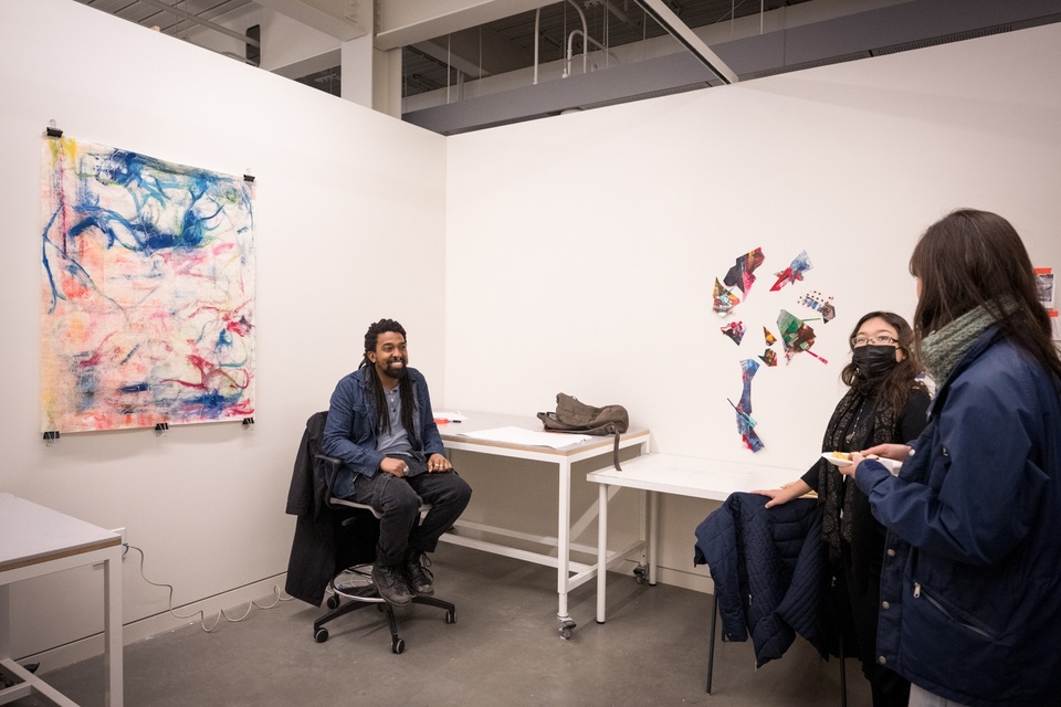 Student seated in a rolling chair smiles at two visitors. A square canvas is clipped to the wall with binder clips behind the student. Dry-brushed textures and lines are painted on the canvas in bright colors.