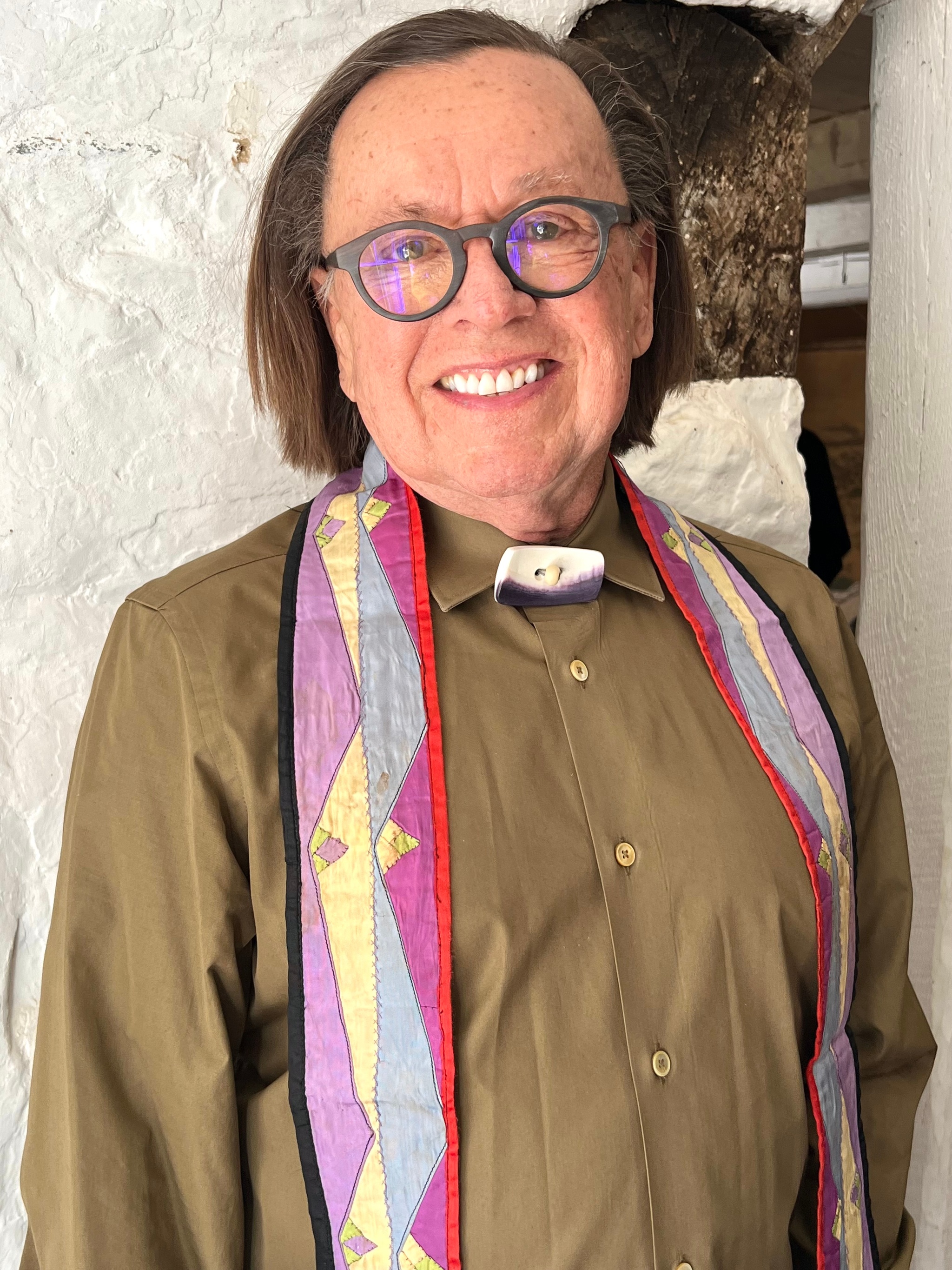 A portrait of Joe Baker. He smiles at us standing with arms at his side. He has chin-length hair parted to one side and wears glasses. He wears an olive-green button down shirt with a narrow stole draped around his neck. It has a geometric pattern in pink, yellow, light blue, and magenta. 