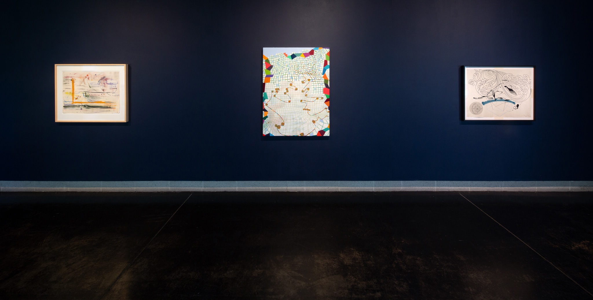 A dimly lit room with three abstract artworks hanging on a dark blue wall.
