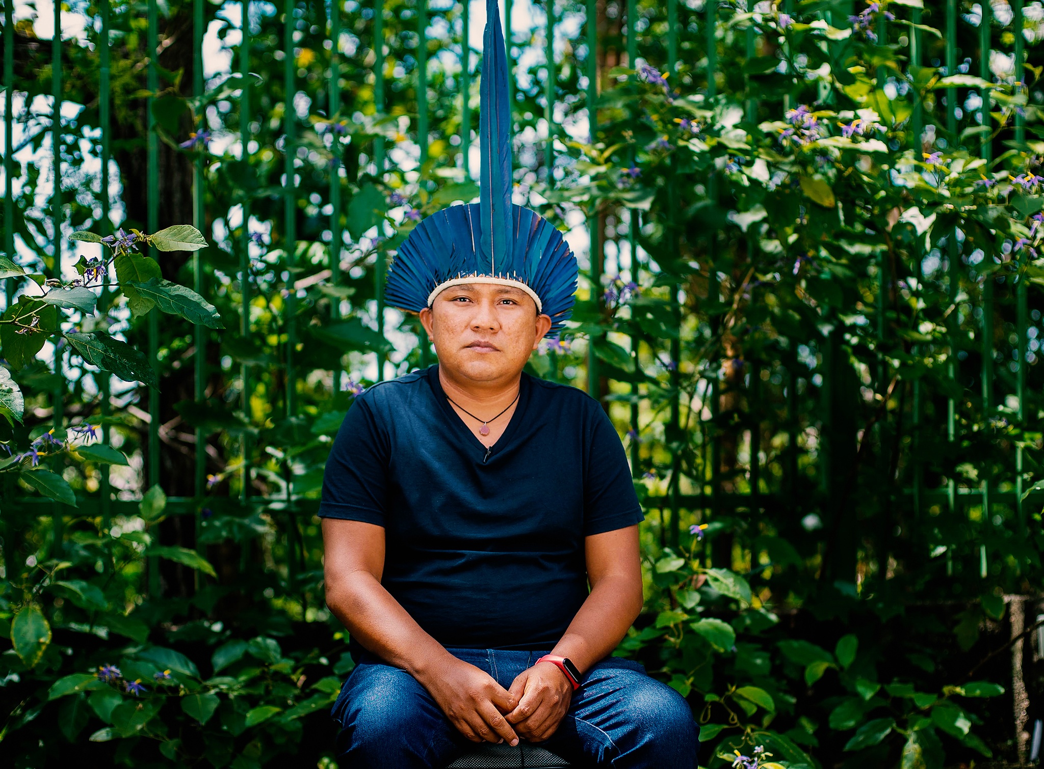 A portrait of Dário Kopenawa. An Indigenous Yanomami man sitting with lush trees behind him. He wears a blue v-neck t-shirt,, blue jeans, and an array of blue feathers on his head.