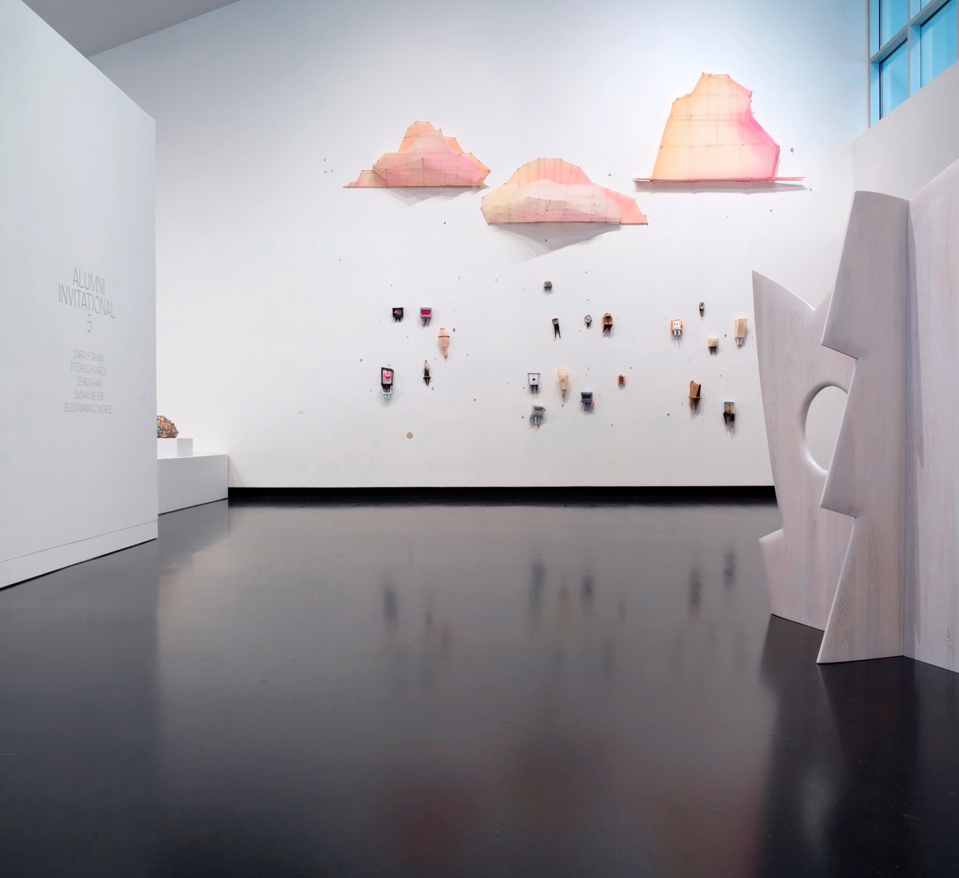 Displayed in a museum gallery is a white, jagged, geometric sculpture in front of many small sculptures that hang on a white wall below pink sculptural clouds.