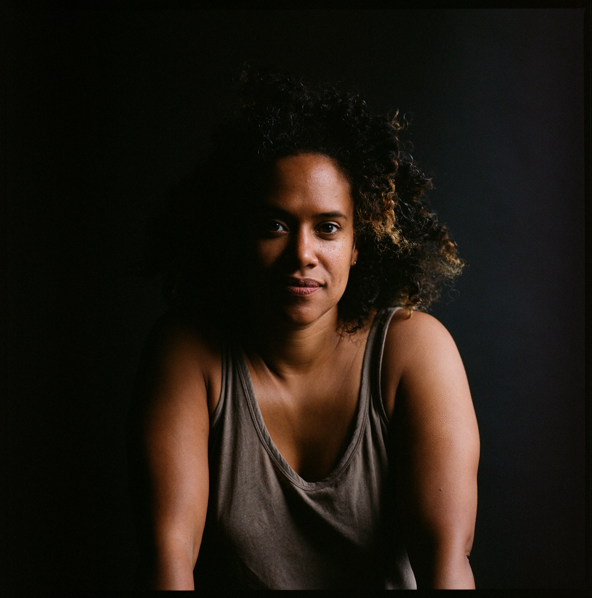 A photo of the artist Leslie Cuyjet who wears a loose tank top and sits against a dark background with her face partly in shadow.
