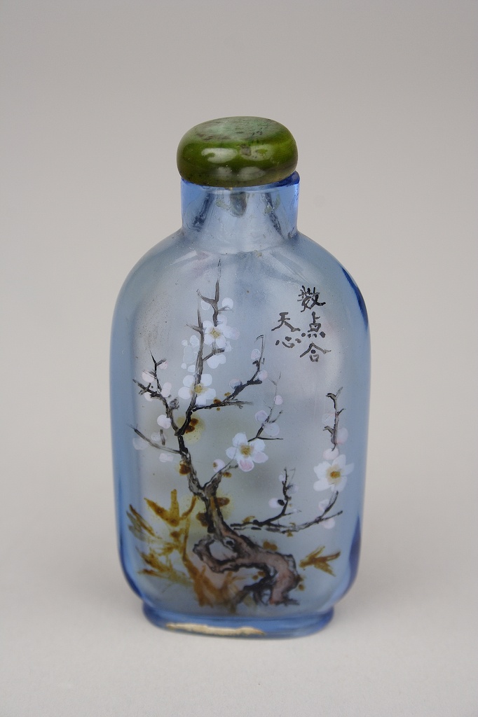 A light blue glass bottle decorated with painting of a flowering tree and Chinese lettering with green stone stopper on top.