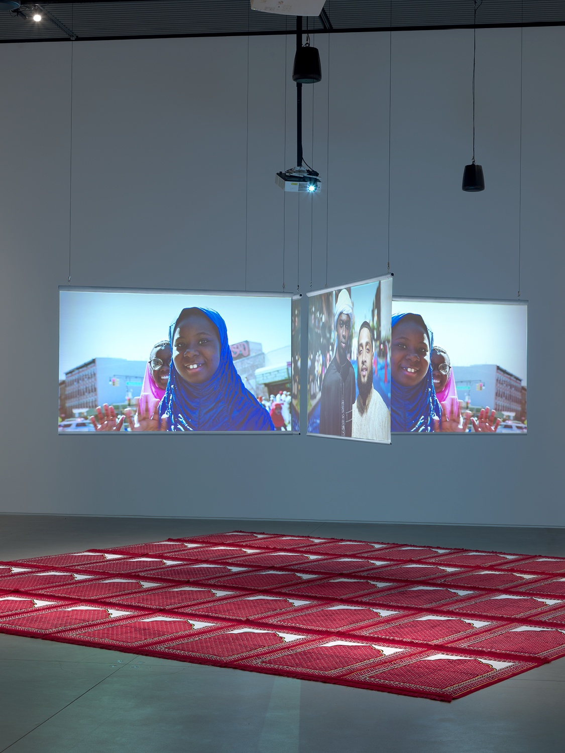 Three video screens suspended in an intersecting formation above a floor covered with red prayer mats placed side by side to form one large square. The screens show a young girl in a headscarf smiling at the camera.