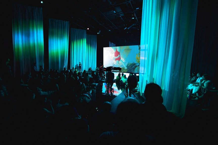 Audience viewing film on large screen in blue-lit space.