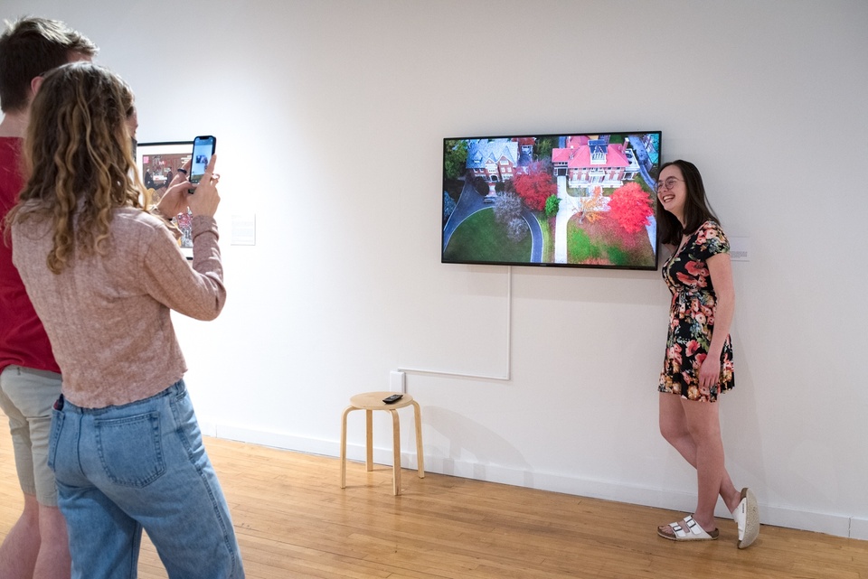 Person stands next to a video screen on a gallery wall and smiles for a photo. On screen is a drone image of a neighborhood.