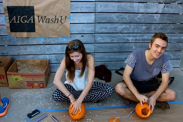 Two students carving pumpkins in front of a wall. The AIGA WashU logo on brown paper hangs from the wall.
