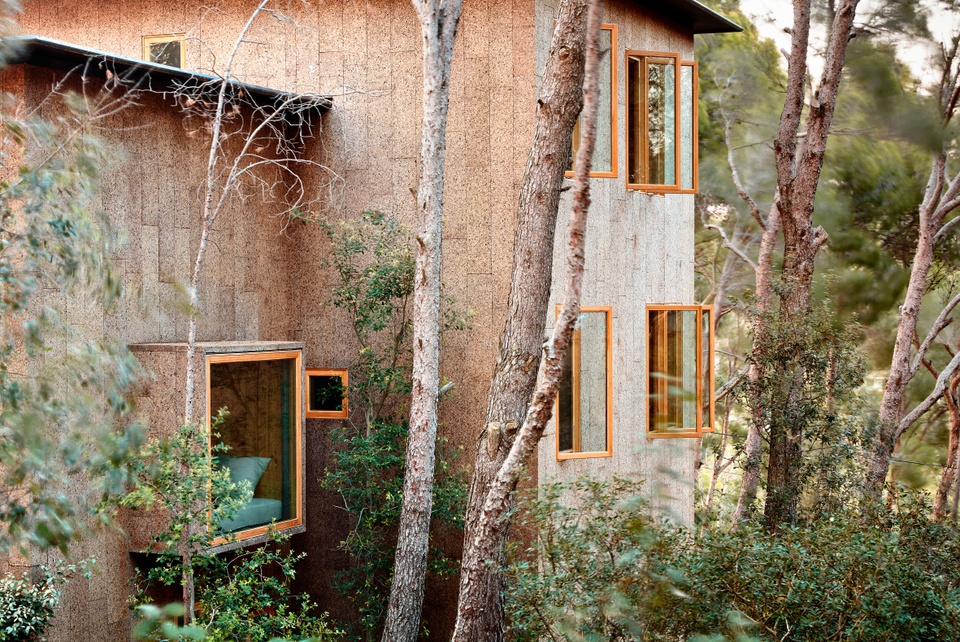 Photo of a multistory house, partly obscured by trees. The house is clad in a light-colored cork material and has two windows, stacked vertically.