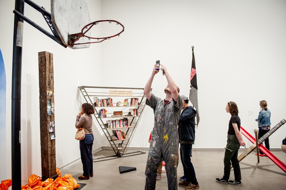 Gallery visitor in a paint-spattered coverall looks up at a basketball hoop in one installation and takes a photo.
