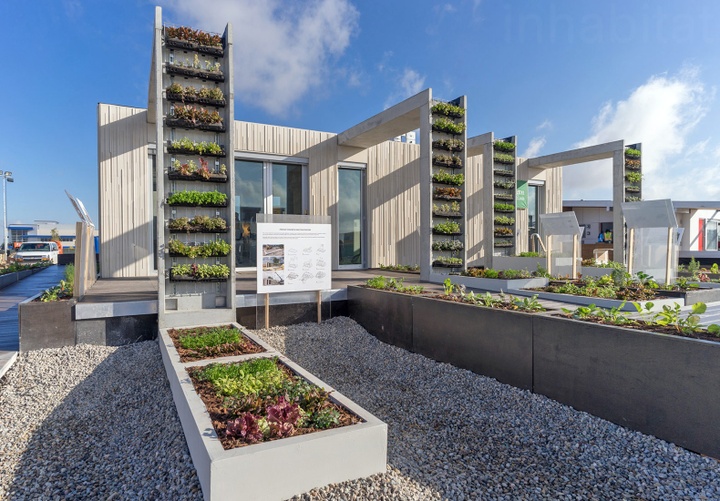 Exterior of CRETE House, a residence made almost entirely from pre-cast concrete. Four L-Shaped structures protrude out from the exterior wall, featuring rows of plants growing. On the ground in front of the house are also several pre-cast concrete planter beds filled with green and red-leaf foliage.