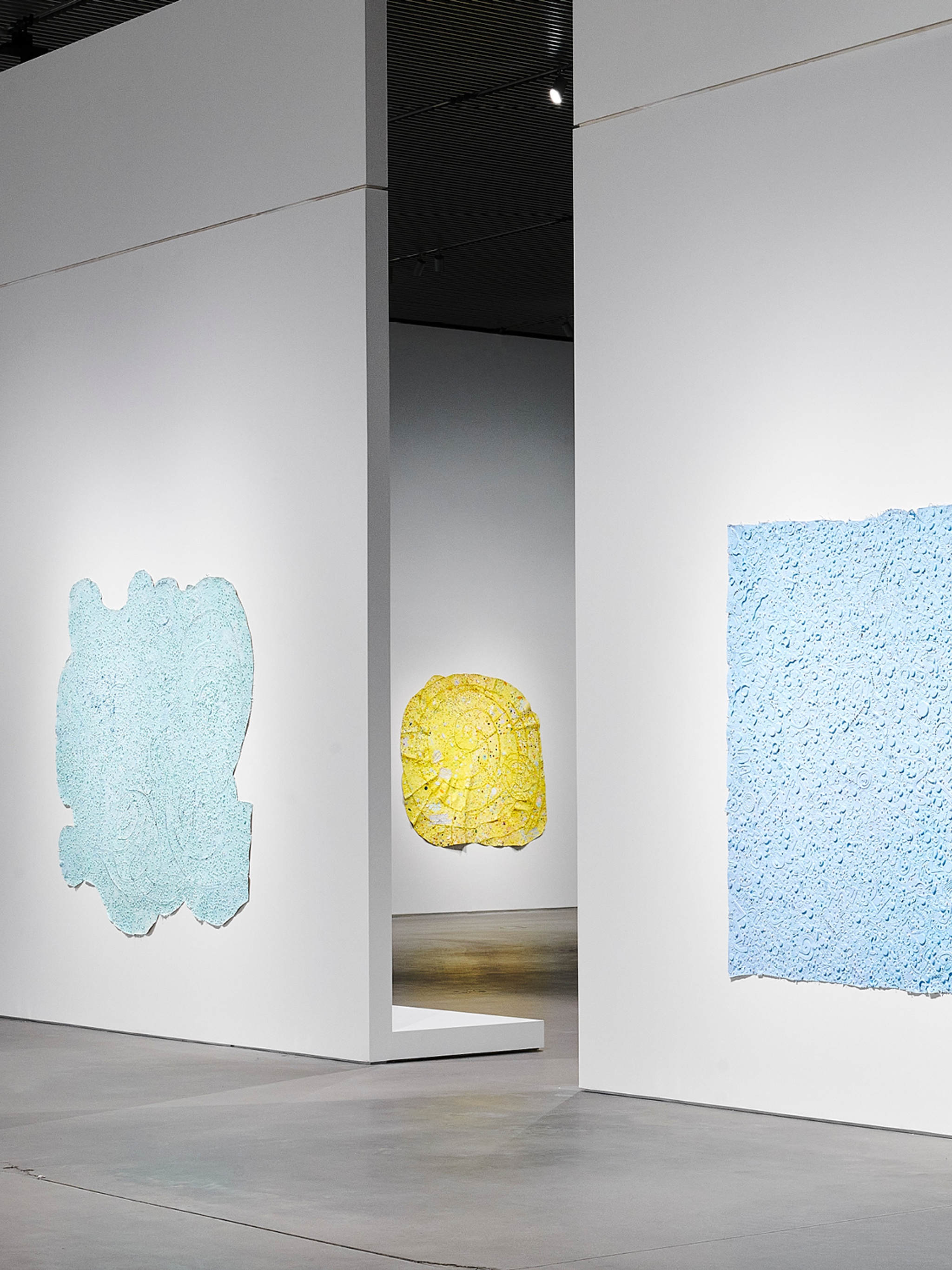 Abstract paintings in the exhibition "Howardena Pindell: Rope/Fire/Water." In the foreground are two blue paintings, with a curvilinear yellow painting visible through a gap in the wall in the background.