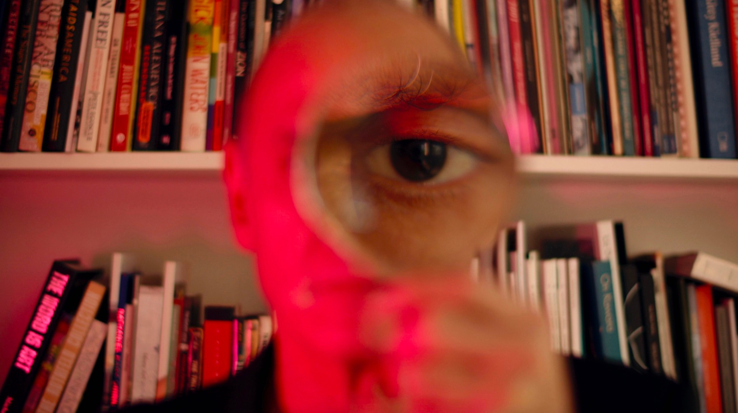 A person in front of a bookshelf holding a magnifying glass, their eye magnified. Red light from the left illuminates them.