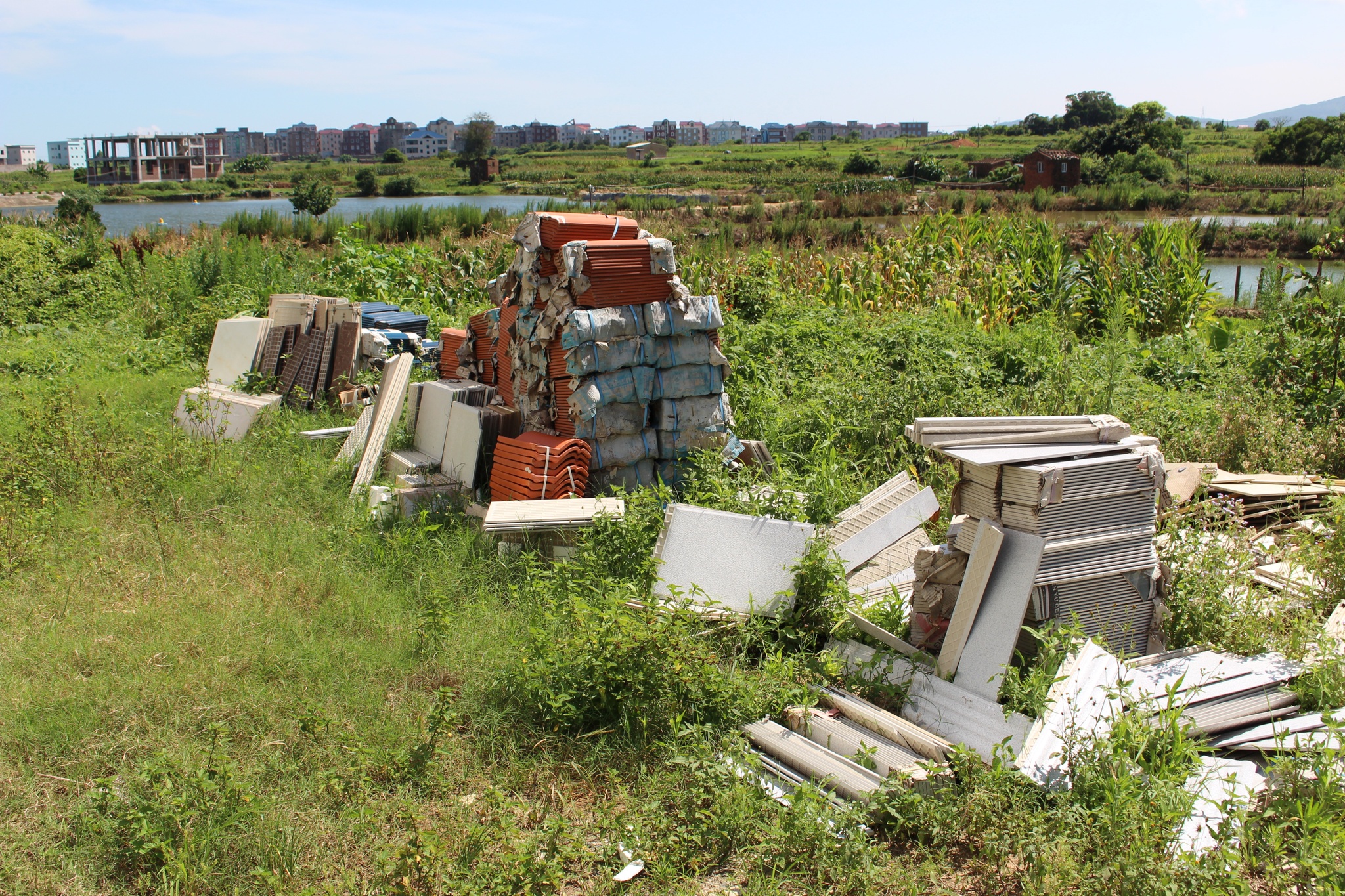 Concrete and terracotta slabs sit in discarded piles in a deserted field or empty lot with a pond and housing development in the distance behind them