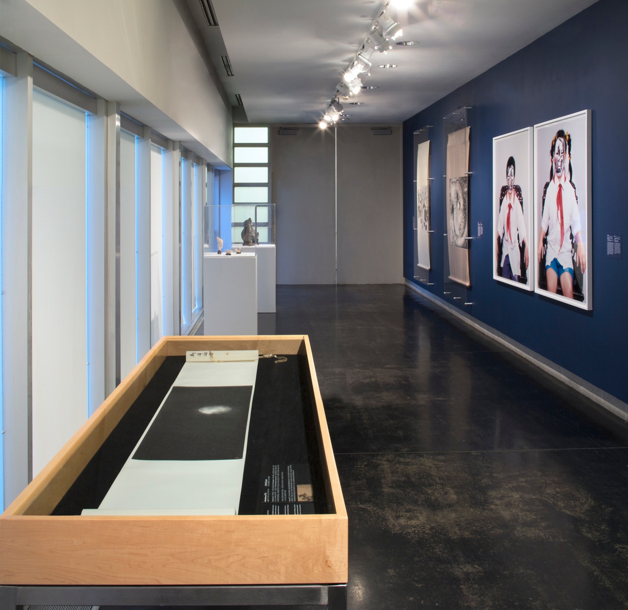 A long, narrow gallery with a case in the left forefront and pedestals behind. Scrolls and framed work hang on a wall painted dark blue on the right.