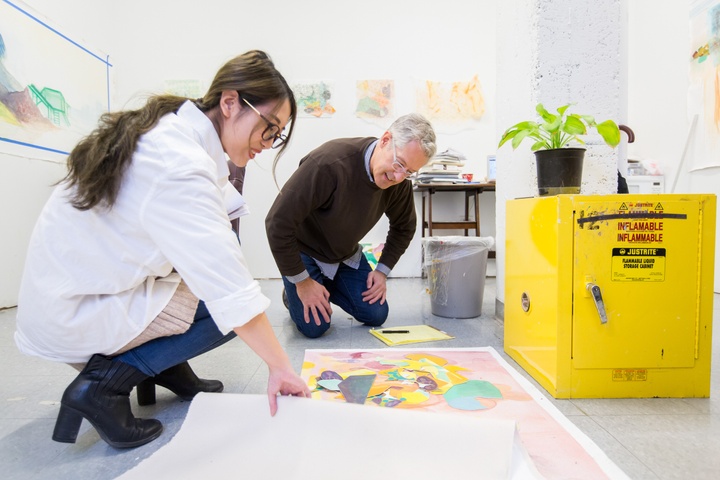 Two people crouch over a large sheet of paper with collaged pieces on the floor of an artist's studio.